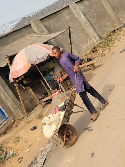 The image portrays an outdoor scene in Puerto López, Colombia, under bright daylight. A young adult male is depicted in motion, pushing a metal wheelbarrow loaded with a large, light-colored tied sack and a dark object. The wheelbarrow's side has "RIB" marked on it. The male wears a striped purple tunic, dark trousers, and light-colored sandals. To his left, a makeshift market stall is visible, constructed with wooden supports and shaded by a partially open, multi-striped umbrella (white, beige, and orange/red). The stall displays various fruits, including green, orange/brown, and yellow/green items, on a wooden surface. The background features a light grey concrete wall topped with barbed wire. The ground is an uneven, dry dirt or sandy surface with scattered debris, including a single dark shoe. A wooden bench is visible to the right of the stall.