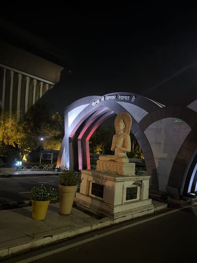The image depicts a nighttime scene in Noida, India, featuring a large arched gateway with a statue of the Buddha at its center. The gateway is illuminated with white and red lights, creating a dramatic effect against the dark sky. Above the arch, text in Hindi reads "नमो बुद्धाय आपका स्वागत है" which translates to "Namo Buddhaya, welcome." The Buddha statue is seated on a pedestal and is made of stone. To the left of the gateway, trees are visible, also illuminated by artificial lighting, casting long shadows. In the foreground, two potted plants are placed on a concrete platform. The scene appears to be a public area, possibly a park or a monument entrance. The overall ambiance is serene and spiritual, enhanced by the nighttime lighting.