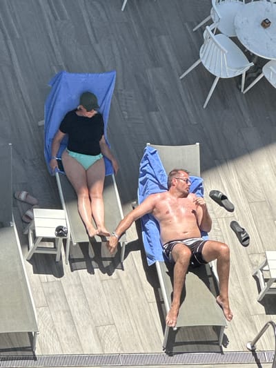 The image shows an overhead view of two people relaxing on lounge chairs by a pool in Lloret de Mar, Spain. On the left, a person wearing a black t-shirt and light blue bikini bottoms is lying on a lounge chair covered with a blue towel. On the right, a shirtless man wearing black and white striped swim trunks and sunglasses is reclining on another lounge chair, also covered with a blue towel. Two black flip-flops are in the air between the lounge chairs, suggesting they were thrown. The setting appears to be a hotel or resort area, with a tiled patio floor and lounge chairs. The image is taken from a high angle, looking down on the scene.
