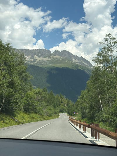 The image depicts a scenic mountain road in Vallorcine, France. The road curves gently upwards, bordered by a wooden guardrail and lush green trees on either side. In the distance, a majestic mountain range dominates the skyline, with its peaks partially obscured by dramatic clouds. The sky is a vibrant blue, interspersed with fluffy white cumulus clouds. A few cars are visible on the road in the distance, suggesting a mild level of traffic. The environment appears to be a natural, undeveloped area, characteristic of a mountainous region. The presence of a road and guardrail indicates it is a passable route for vehicles.