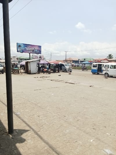 The scene is a street corner in Ovwian, Nigeria, a town in Delta State. It is a sunny day, with a few clouds scattered in the blue sky. In the background, there are several buildings with flat roofs. The street is paved with concrete blocks, and the sidewalk is cobblestone. There are several vehicles, including a white van, a blue van, and a motorcycle, parked along the street. There is a large billboard advertising a company called "Partner Tech." A few people are standing on the sidewalk, and some are sitting on a bench. There are also a few small shops and kiosks on the street.