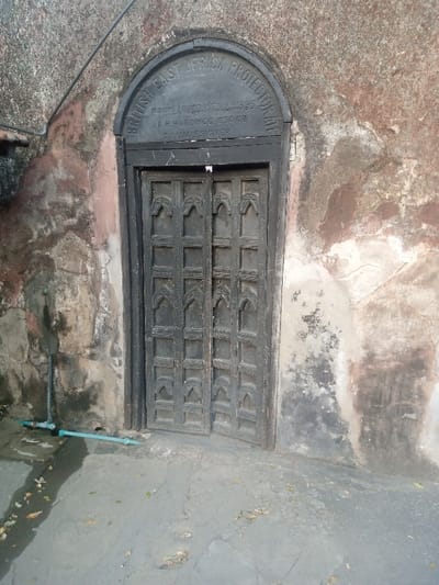 The image displays a dark, weathered, double-leaf wooden door set within a decaying, rough-textured wall. Above the door, an arched stone plaque is inscribed with "BRITISH EAST AFRICA PROTECTORATE PROCLAIMED 1ST JULY 1895 A.H. HARDINGE ESQR. COMMISSIONER." The door features multiple carved rectangular panels, each with an arched decorative motif. The surrounding wall, predominantly reddish-brown, grey, and off-white, shows significant peeling, staining, and material degradation. To the left of the door, a thin, dark cable descends diagonally from the top left corner. Near the base of the left wall, two light-blue plastic pipes are visible, one running vertically and the other horizontally along the ground, connecting at an elbow joint. The ground directly in front of the door is a grey, uneven paved surface with scattered dry leaves and dark stains, particularly near the pipes. No people are present. This scene is located in the Makadara/Mji Wa Kale ward, Kenya, consistent with the historical marker and architectural style of Mombasa's Old Town.