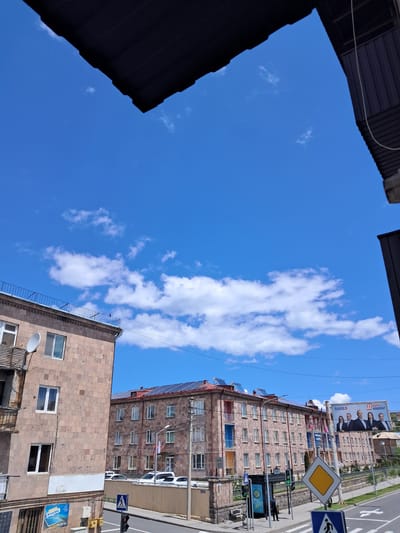 The image presents a vibrant urban scene on a sunny day, captured from a low vantage point looking upwards. A vast expanse of clear blue sky, dotted with scattered white cumulus clouds, dominates the upper and left portions of the frame. Dark, angular architectural features, possibly parts of roofs or awnings, frame the top-left and bottom-left corners, creating a visual enclosure for the sky.
On the right side of the image, multiple multi-story buildings constructed from light brown or tan brick/stone line a street. These buildings feature numerous rectangular windows, many with white frames. Solar panels are visible on the roof of one building, suggesting a focus on renewable energy. A white satellite dish is also attached to the facade of a building closer to the viewer.
A street runs alongside the buildings, with several white cars parked along its edge and visible road markings. Prominent street signs include a yellow diamond-shaped sign, indicating priority or a main road, and a blue rectangular sign with a white pedestrian icon, signifying a pedestrian crossing. A traffic light or lamppost stands near these signs.
A large billboard is mounted high on one of the buildings, displaying a group of four men in suits. Visible text on this billboard includes "AT" and Armenian script, with legible words like "ԱՆԴՐԵԱՍ" (Andreas) and "ՆԱՐԵԿ" (Narek), suggesting a political or public figure advertisement. Smaller signs on the buildings display "AYSOLAR" and "ԷՆԵՐԳԵԼ" (Energell) alongside a phone number "098 555 555", likely advertising local businesses related to solar solutions and energy. The bright light and distinct shadows indicate it is daytime.