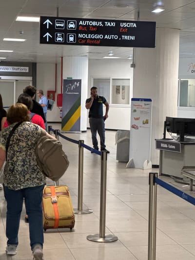 The image shows the interior of an airport in Cinisi, Italy, with people standing in a queue near the customs area. A man in uniform is standing by, speaking on a mobile phone. In the foreground, people are waiting with luggage. Signage indicates directions for "AUTOBUS, TAXI" and "NOLEGGIO AUTO, TRENI", meaning bus, taxi and car rental, trains. A sign reads "DOGANA-Customs" indicating the customs area of the airport. Another sign describes travelling with €10,000 or more.