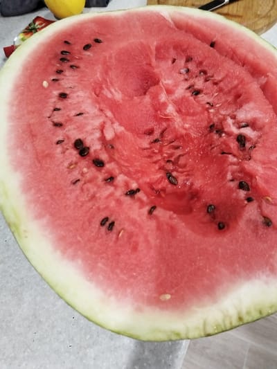 A close-up shot captures a ripe, red watermelon cut in half, revealing its juicy interior and scattered black seeds. The watermelon is positioned on a speckled gray surface, possibly a countertop or table, with a glimpse of a wooden cutting board and a knife in the background. A yellow object, likely a lemon, is also visible behind the watermelon. The lighting suggests an indoor setting, possibly a kitchen. The image focuses on the texture and color of the watermelon, emphasizing its freshness. There is no visible text in the image. The scene appears to be set in Bezlesnyy, Russia.