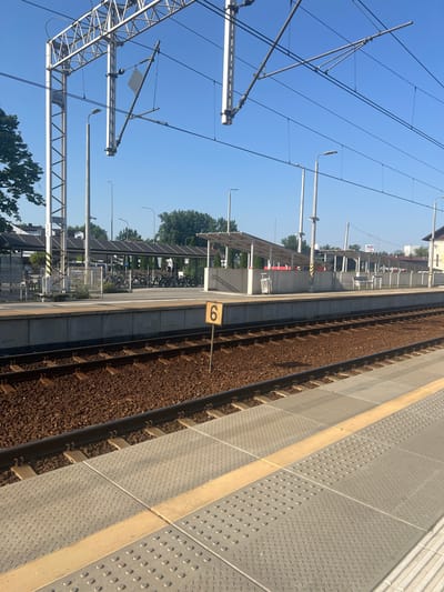 This is a daytime, eye-level, medium shot of a train station platform in Skawina, Poland. The foreground is dominated by a train track, with ballast and railway sleepers visible. To the right of the track is a textured gray platform with tactile paving and a yellow warning strip. A yellow sign with the number "6" is positioned on the platform, facing the camera. In the middle ground, behind the platform, is a covered bicycle parking area with numerous bicycles parked under a white, transparent roof supported by pillars. Across from the platform, a modern building with a white facade and a dark roof is visible in the background, along with trees and utility poles. Overhead, power lines for electric trains are suspended from a metal framework. The sky is clear and blue.