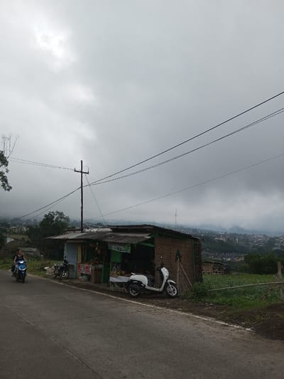 The image displays a roadside scene in Ambakirano, Madagascar, under a heavily overcast, grey sky. A paved road extends from the lower left to the right foreground, curving slightly.
In the mid-ground, on the left side of the road, a person wearing a dark top rides a blue scooter, traveling from left to right. Adjacent to the road on the right, a single-story, rustic roadside shop with a corrugated metal roof is visible. The shop's facade features sections of light blue and green paint, and merchandise, including what appears to be coconuts or melons in sacks and hanging items, is displayed at the front. A dark motorcycle is parked in front of the shop, and a white scooter is parked further to its right. At least one individual is partially visible inside the shop, obscured by the displayed goods.
A dark utility pole with a horizontal crossarm stands just to the left of the shop, supporting multiple overhead power or communication lines that extend across the upper portion of the frame. To the right of the shop, a simple wooden fence borders a green, cultivated area. The background features a sprawling settlement of buildings across hillsides, partially obscured by a thick layer of clouds or mist. A distant communication tower is discernible on a higher peak in the hazy background.