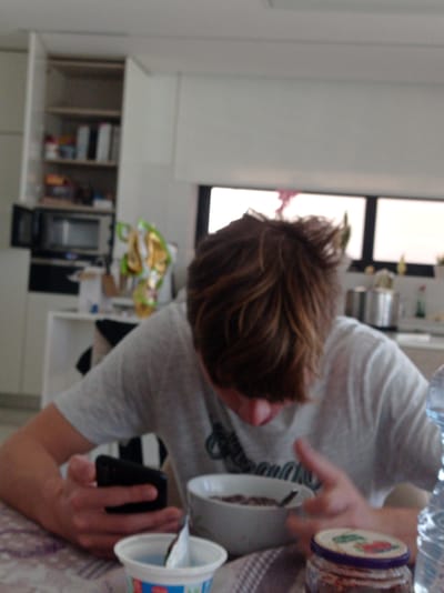A person is seen indoors, possibly in a kitchen setting, holding a phone in one hand and two yogurt containers. A jar is also in the scene. The person is wearing a gray shirt. There's a window visible in the background and kitchen cabinets and appliances. No specific information suggests a location in Felgueiras, Portugal. No action or interaction is clearly taking place.