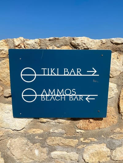 A blue sign with white text is attached to a rough stone wall under a clear blue sky. The sign has two directional indicators. The top line reads "TIKI BAR" with an arrow pointing to the right. The bottom line reads "AMMOS BEACH BAR" with an arrow pointing to the left. The sign is mounted on a wall made of irregular, beige and gray stones. The location is Panormos, Greece.