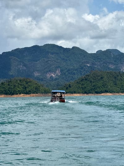 The image shows a boat moving on the water with mountains in the background. The boat is red and has a blue canopy, and there are several people on board. The water is turquoise and choppy, and the mountains are covered in lush green trees. The sky is partly cloudy. The scene is likely Khao Sok National Park, near Ban Ta Khun, Thailand, known for its stunning natural beauty and limestone karsts.