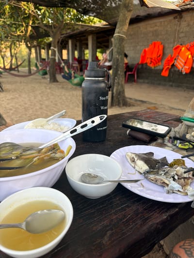 In an outdoor setting, likely a restaurant or picnic area in Bắc Trạch Commune, Vietnam, a meal is being served on a dark wooden table. The scene is casual and appears to be during the daytime, judging by the natural light.

Dominating the table are several white plates and bowls. One plate holds a partially eaten fish, its head and bones visible. Adjacent to it are two white bowls; one contains a portion of rice, and the other holds a clear soup with visible ingredients. Another white bowl sits nearby, empty except for a spoon. A silver fork rests on the rim of the soup bowl.

On the table, to the left, is a black water bottle with white text that reads "Stay POWRFUL Work". Further left, two bright orange life vests are hanging, suggesting the proximity to water or an activity involving water. A smartphone with a black case is also present on the table, leaning against a small brown object.

In the background, out of focus, there are glimpses of trees and what appears to be a structured seating area with people in the distance. The overall impression is a relaxed, informal meal enjoyed outdoors.