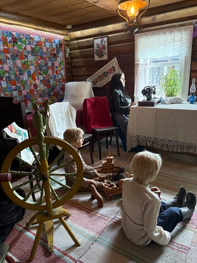 The image depicts a dimly lit interior of what appears to be a historical dwelling, possibly a museum exhibit in the village of Ovsjanka, Russia. The room has log walls and a wooden ceiling. In the foreground, two mannequins are positioned on a colorful striped rug. One mannequin, facing away from the camera, is a child with blond hair wearing a white shirt and dark trousers, seated with legs extended. Another mannequin, partially visible to the left, is also seated and appears to be a child with blond hair, wearing a patterned shirt. A prominent yellow and green spinning wheel stands to the left of the frame. In the background, a woman mannequin, dressed in traditional dark clothing and a headscarf, is seated at a table covered with a white lace tablecloth. On the table, a black sewing machine and a blue kerosene lamp are visible. A large window with white curtains is behind the table, and a plant grows in a pot on the windowsill. A colorful patchwork quilt hangs on the wall to the left, and a framed picture of flowers is on the wall above the woman. A red garment is draped over a chair. The scene is illuminated by a hanging oil lamp with a warm glow.