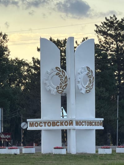 The image shows a sign in Mostovskoy, Russia. The sign is white with gold lettering and features two circular emblems, each with a gear-like design surrounded by laurel leaves. The text on the sign reads "МОСТОВСКОЙ". The sign is positioned outdoors, with trees and a cloudy sky visible in the background. The lighting suggests it is daytime, possibly late afternoon or early evening given the soft light. There are some white, segmented barriers with red flowers visible to the right of the main sign, and a road or pathway beyond them. A small red sign with white text, reading "Кафе", is visible below the main sign. The scene appears to be a roadside entrance or landmark.