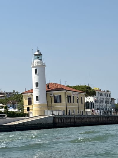 An image shows a lighthouse and a yellow building with a red-tiled roof in Cesenatico, Italy. The lighthouse is white and cylindrical with a lantern room at the top. The building appears to be the Guardia Costiera, an Italian coast guard station, as suggested by the text on the building. Several people are visible on the promenade in front of the buildings, and a body of water, likely the Adriatic Sea, occupies the foreground. The sky is clear and blue.