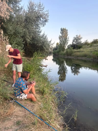 Two men are fishing on the bank of a calm river with dense vegetation on both sides. The man in the foreground, wearing a colorful short-sleeved shirt with a pattern of red and yellow shapes and patterned shorts, is sitting down and holding a fishing rod. The man in the background, standing and wearing a maroon t-shirt, light-colored shorts, and a beige cap, appears to be attending to his fishing line. The water reflects the trees and the clear sky above. The overall setting suggests a natural, tranquil environment on the outskirts of Krasnoflotskoye Rural Settlement, Ukraine.