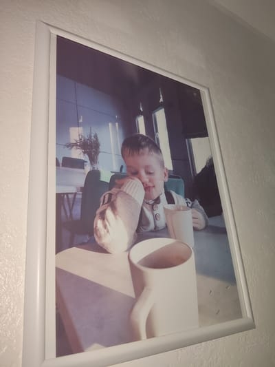 A framed photograph hangs on a textured wall, depicting a young boy with his eyes closed, leaning on his arm. He is wearing a light-colored sweater with decorative buttons and a knitted item on his arm. In front of him are two white mugs, one partially obscured by the other. The background suggests an indoor setting with tables and chairs, possibly a cafe or restaurant. The lighting creates shadows on the table. The location of Reftinsky, Russia is not discernible from the image.