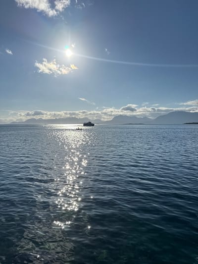 A wide-angle, horizontal, exterior shot captures a sun-drenched body of water with a distant mountain range under a bright blue sky. The sun is positioned in the upper left quadrant of the frame, with bright rays beaming outward. A faint halo effect is visible around the sun. A few scattered white clouds are present in the sky. The water in the foreground is calm with a slight ripple, reflecting the sunlight in a glittering path that stretches towards the horizon. A small boat with what appear to be a few people on it is visible in the mid-ground, situated on the sunlit path on the water. Further in the distance, a larger ferry or ship is moving away from the viewer towards the right side of the frame, also on the reflective water. The background features a long, silhouetted mountain range, likely part of the Norwegian landscape near Harstad, under a hazy sky. The lower portion of the water appears darker and clearer, with some submerged rocks or seabed visible through the surface. The scene depicts a tranquil day at sea, likely in the vicinity of Harstad, Norway, with clear skies and calm waters.