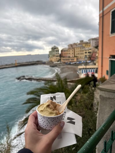 A right hand, displaying light purple nail polish, holds a white paper cup of gelato in the lower-center of the frame. The cup features a black polka-dot pattern and contains two scoops of gelato, one light green and one light brown, with a light-colored wooden spoon inserted. Two white paper napkins, branded with "NUGHE" and a stylized 'N' in black, are partially held beneath the cup. The subject's dark sleeve is visible on the lower left. To the lower right, a dark green metal railing and a light-colored concrete ledge are present.
The background, viewed from an elevated vantage point, depicts the coastal town of Bogliasco, Italy, under an overcast sky filled with grey clouds. The turquoise sea, exhibiting small waves, occupies the left side of the frame. A dark, concrete pier with a yellow navigational beacon extends into the water from the left. A pebbly beach, populated by several small boats pulled ashore, curves along the coastline to the right of the pier. Beyond the beach, a cluster of multi-story buildings in characteristic Ligurian pastel shades of yellow, orange, and light green ascend the hillside. Blue and white striped structures, likely beach amenities, are visible near the beach. A salmon-pink building with a dark green shuttered window is partially visible on the far right. Several cargo ships are discernible on the distant horizon line where the sea meets the sky. Lush green foliage, including palm fronds, is visible in the midground, between the foreground subject and the distant town.