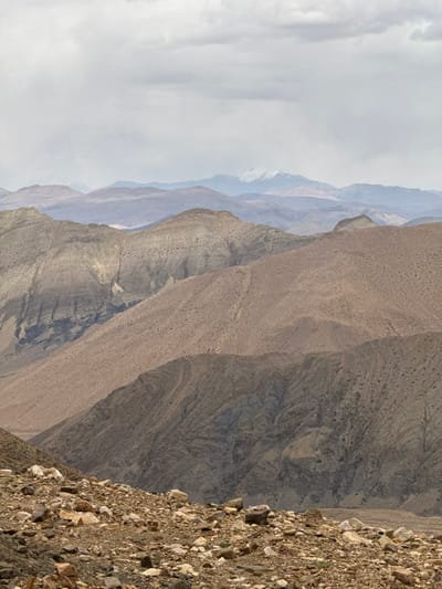 The image depicts a mountainous landscape, with layered rock formations and varying terrain. In the foreground, a steep slope is covered with loose rocks and gravel. The middle ground features a series of ridges and valleys, showing exposed rock faces with different textures and patterns. In the background, more distant mountain ranges are visible, fading into a hazy sky. There are no people visible in the frame. The location is likely a remote, rocky area, but the precise location cannot be determined from the image alone.