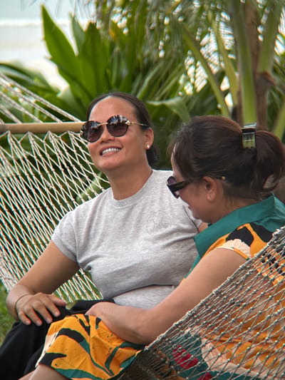 Two women are relaxing in a hammock outdoors, surrounded by lush green foliage and palm trees. The woman on the left, wearing sunglasses and a grey t-shirt, is smiling and looking to her right. The woman on the right, also wearing sunglasses and a colorful patterned shirt, is looking down and to her left. The hammock is made of white rope and is suspended between two points. The background suggests a tropical or resort-like setting, potentially in Dong Ninh Hoa, Vietnam, a coastal town known for its beaches and natural beauty.
