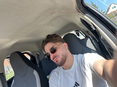 A man is taking a selfie inside a car. The car's interior has a light gray roof lining and dark gray fabric seats. The man wears sunglasses and a white t-shirt with an Adidas logo. Through the car window, a house with green shutters and a Swiss flag is visible, suggesting the location is likely Rafz, Switzerland.