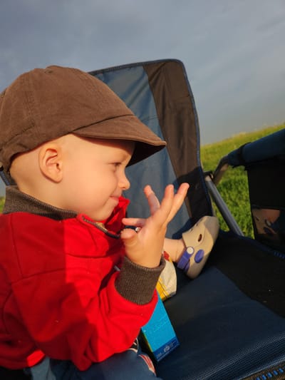 A young child wearing a brown cap and a red jacket sits in a blue camping chair outdoors. The child is looking to the right, with their left hand raised, fingers spread. A small shoe with purple and white accents is visible on their foot. A blue box with text is positioned near the child's leg. The background consists of a grassy field under a cloudy sky. The image appears to have been taken in Atameken, Kazakhstan.