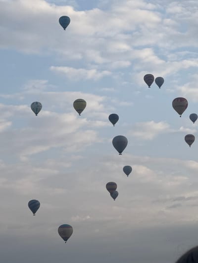 The image shows a sky filled with numerous hot air balloons, suggesting the location is likely the popular ballooning destination of Göreme, Turkey. The balloons vary in color and design, some striped and some with lettering. They are dispersed across the sky, surrounded by a backdrop of white and gray clouds. The sky itself appears to be partly cloudy, with patches of blue peeking through. The angle of the photograph suggests it was taken from below, looking up at the balloons.