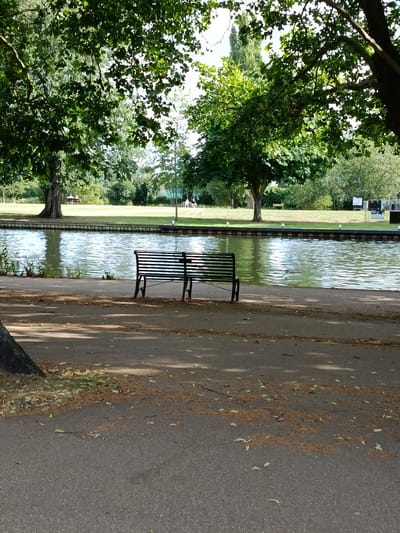 A sunny day in a park in Bedford, United Kingdom, with a canal in the foreground. A black metal bench sits on a paved path, facing the water. The canal reflects the bright blue sky and the lush green trees that line its banks. Further back, a grassy field stretches towards a line of trees. Two people are seated on a bench in the distance, enjoying the serene atmosphere. In the background, there are some signs and structures, possibly part of a recreational area. The overall mood is peaceful and idyllic, suggesting a perfect day for outdoor relaxation.