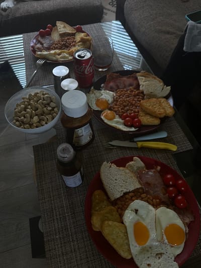 This image depicts an indoor scene in Dorchester, United Kingdom, focusing on a dark-surfaced coffee table presenting three individual breakfast servings.
The top-left plate, reddish-brown, holds bacon, baked beans, two slices of toast, and five cherry tomatoes. A silver fork is positioned on this plate. The middle-right plate, brown, contains bacon, baked beans, two slices of white bread, two fried eggs with intact yolks (sunny-side up), five cherry tomatoes, and two hash browns. The bottom-right plate, red, displays bacon, baked beans, two slices of white bread, two fried eggs with intact yolks (sunny-side up), four cherry tomatoes, and three hash browns. A silver fork and a knife with a yellow handle are placed beside this bottom-right plate.
To the left of the table, a white bowl is filled with unshelled cashew nuts. Adjacent to the bowl are two silver-colored shakers, likely for salt and pepper. An open red Coca-Cola can and an empty, clear glass tumbler are situated near the center of the table. Two brown condiment bottles are also present: a larger one with a white cap and a smaller one with a darker cap.
In the background, a light-colored sofa with a dark throw is visible. A partial view of an animal, consistent with a domestic dog, rests on the sofa in the upper left corner. Natural light, entering from a window with horizontal blinds, casts distinct striped patterns across the table surface and the light-colored floor. No human figures are visible.