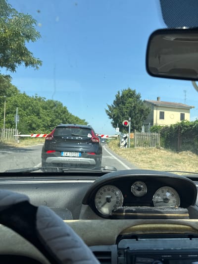 The image is taken from the driver's perspective inside a car, looking through the windshield. The car appears to be stopped at a railway crossing, indicated by the lowered red and white barrier across the road. Ahead of the car, a black Volvo SUV with license plate "EW 590YD" is visible, also stopped. To the right of the Volvo, a traffic signal displays a solid red light, further confirming the stop at the crossing. Beyond the crossing, a rural landscape with trees and a yellow house can be seen. The interior of the car shows a dashboard with various gauges, including a speedometer and a tachometer, and a steering wheel cover. The overall scene suggests a sunny day. The presence of Italian license plates on the Volvo and the distinctive architecture of the house point to a location in Italy. Specifically, the details in the image are consistent with a railway crossing in or near Casalmaggiore, Italy, a town known for its rural setting and proximity to railway lines.