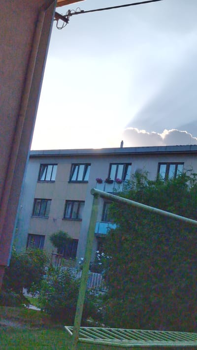 This media file, captured in Vysoké Mýto, Czechia, depicts a building with multiple windows, some adorned with flower boxes. A dense cluster of trees partially obscures the building on the right. A section of a balcony railing, painted green, is visible in the foreground, along with what appears to be another roofline. The sky is bright, with a large cloud formation and visible sunbeams radiating outwards. Electrical wires are also present in the sky. The image is oriented vertically, suggesting it may have been taken from a slightly elevated perspective or a mobile device held sideways. The lighting indicates it is daytime, likely late afternoon due to the strong contrast and sunbeams. There is no visible text.