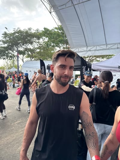 A medium shot captures a man with dark hair and a beard, wearing a black t-shirt with "FITPRO" printed on it. He is looking towards the camera with a slight smile. A portion of a tattoo is visible on his right arm. Behind him, there are tents, people, and trees. The setting appears to be a gathering or event with individuals performing exercises, possibly a fitness event in บ้านสำนักตะแบก, Thailand. Some are doing push-ups. A woman with a tattoo on her arm is also in the shot.