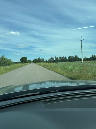 The image depicts a view from a vehicle traveling along a paved road in a rural setting. The road stretches ahead, bordered by a grassy field on the left and trees in the distance. Overhead, a blue sky is visible with scattered white clouds and a faint contrail from an aircraft. A telephone pole with wires stands beside the road. The right side of the image shows the interior of the vehicle, including a dark-colored panel with a textured surface and reflections on a glossy surface, likely a window or dashboard. The scene suggests a daytime journey under clear, possibly warm weather conditions. There is no visible text in the image.