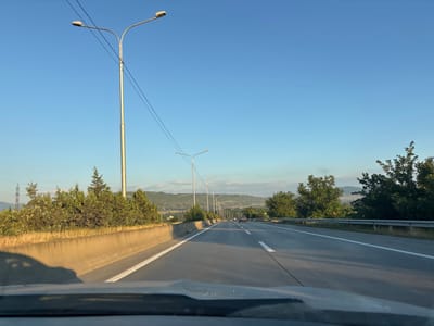 The image depicts a view from a vehicle traveling on a multi-lane highway. The highway is paved with asphalt and marked with white lane dividers. On the left side of the highway, there is a concrete barrier with vegetation and trees behind it. The background shows rolling hills and mountains under a clear blue sky. Several lampposts line the side of the highway. In the distance, a few cars can be seen on the road. The time of day appears to be late afternoon or early evening, given the warm light and the long shadows. This scene is consistent with the landscape found along the highway leading to Dzveli Kanda, Georgia, characterized by its mountainous terrain and roadside infrastructure.