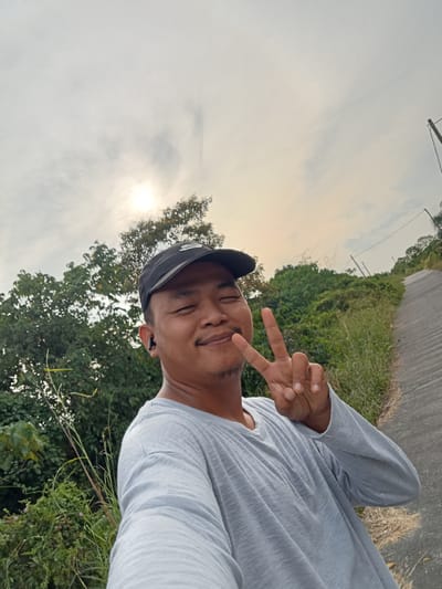A man wearing a black baseball cap and a light gray long-sleeved t-shirt is posing for a selfie outdoors. He is smiling and making a peace sign with his right hand. He has a small mustache and goatee and is wearing a black wireless earbud in his left ear. Behind him, there is lush green vegetation and trees. A paved path or road slopes upwards to the right, with power lines visible in the distance. The sky is overcast with the sun shining through the clouds. The overall scene suggests a rural or semi-rural setting, possibly on the outskirts of Teluk Kenidai, Indonesia.