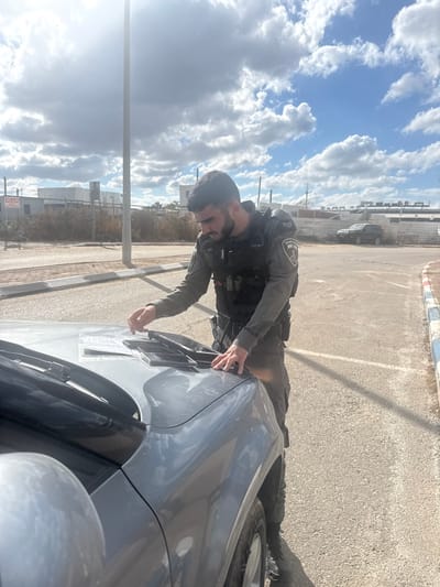 A male individual in a grey tactical uniform, including a black tactical vest with an Israeli Police emblem on the right sleeve and a holstered handgun, stands next to a light grey SUV in Lod, Israel. He possesses a dark beard and is observed manipulating documents and a dark wallet-like object on the vehicle's hood with his right hand, while his left hand rests on the hood. The setting is an outdoor paved area featuring a road with a white and blue painted curb. The background reveals a tall metal pole, a red and white 'No Entry' sign in Hebrew (אסורה כניסה), an unpaved section, low industrial buildings enclosed by a fence, and a dark SUV parked further away. The sky is partly cloudy with direct sunlight visible from the upper left, causing a glare.