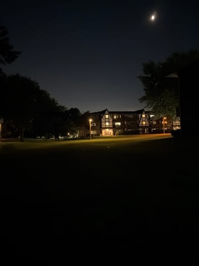 This is a nighttime photograph taken in Vernon Hills, United States of America. The image captures a residential building with lights on, suggesting occupancy. The building appears to be an apartment complex or condominium, characterized by its multi-story structure and multiple windows, some of which are illuminated. The architecture features gabled roofs and what appear to be decorative timber framing.
In front of the building, a well-maintained lawn is visible, illuminated by streetlights that cast pools of yellow light. Several trees are scattered across the scene, their foliage appearing dark and dense against the night sky. The sky itself is dark, with a crescent moon and a single star visible, indicating a clear night. The overall atmosphere is quiet and residential. The foreground is largely consumed by darkness, suggesting the vantage point is from a distance, or perhaps from within a darker area like a yard or another building.
