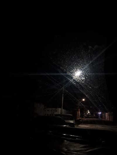 A street scene in Cumana, Venezuela at night. The image is dark, with a bright street lamp in the upper right quadrant illuminating a gritty, textured surface, possibly a window or windshield covered in water droplets or grime. Several power lines crisscross the scene, and another, dimmer street lamp is visible further down the road. The road itself is paved and appears wet. In the foreground, the hood of a vehicle is visible. To the left, a wall with writing on it is partially obscured by darkness, with the words "CRISTO VIENE PRONTO" (Christ is coming soon) discernible. The scene depicts a typical urban street at night, with hints of urban decay and religious messaging.
