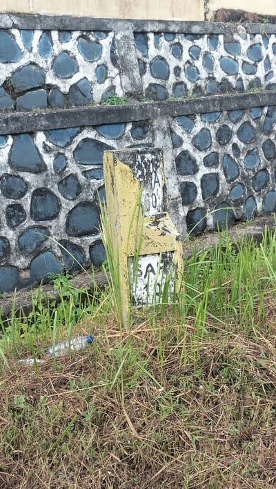 This full-shot image captures an outdoor scene in Lamtamot, Indonesia, featuring a weathered stone wall and a partially broken marker post in the foreground. The wall, likely a retaining wall or barrier, is constructed from dark, irregularly shaped stones embedded in light-colored concrete or mortar, with a prominent horizontal concrete beam dividing its sections.
In front of the wall, a fractured concrete or stone marker stands amidst grass. The marker is predominantly yellow, though much of its paint is chipped and faded, revealing a white or light gray base. Text is partially visible on its surface; the upper fragment shows characters such as "...A 0" or "...IA 0", while the lower part clearly displays "SA" followed by what appears to be a "0" or "O" below it, though the full inscription is unreadable.
The ground around the marker and in the foreground is covered with a mix of green grass and dry, brown vegetation. A discarded clear plastic water bottle with a blue cap lies on the ground amidst the grass to the left of the marker. The scene is illuminated by natural daylight, suggesting it is daytime. There is no visible activity or people, presenting a static view of an aged and somewhat neglected area, possibly roadside or adjacent to a structure.