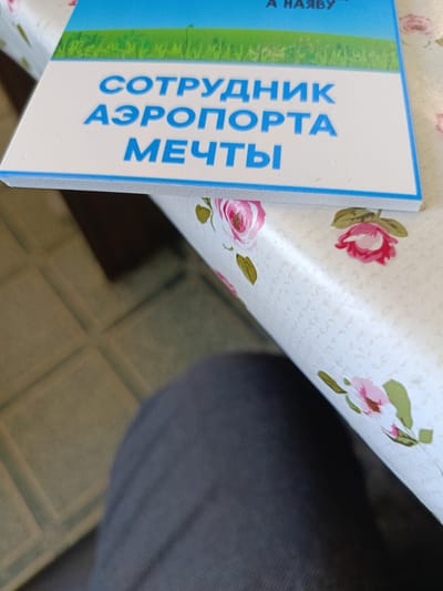 The image shows a close-up of a book with the title "СОТРУДНИК АЭРОПОРТА МЕЧТЫ" (Airport Dream Employee) in blue lettering against a white background. Above the title, a green field and blue sky are depicted, suggesting the cover art. The book is placed on a white tablecloth with a floral pattern of pink roses. In the foreground, a portion of a person's dark grey trousers and a dark blue fabric are visible, indicating the perspective of someone seated and holding or interacting with the book. The background on the left shows a tiled floor in a muted beige and green color. The scene appears to be indoors, likely in a home or office setting. There is no visible indication of the specific location within Semenkovo, Russia.