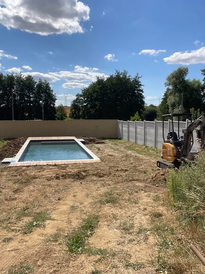 A partially constructed swimming pool sits in a backyard in Viry-Noureuil, France. The rectangular pool is filled with blue water and has a dark-colored liner. The edges of the pool are lined with wood planks, and a grey border surrounds the top. The ground around the pool is bare earth with some scattered dry grass and weeds. A light-colored fence runs along the back of the property, and trees are visible beyond the fence. A small yellow excavator is parked to the right of the pool, with its arm extended. The sky is blue with scattered white clouds.