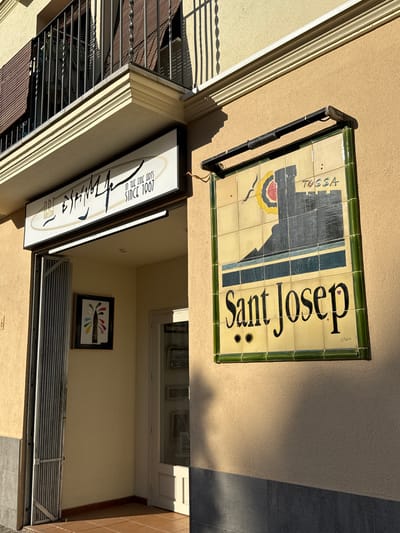 An art gallery, "ART ESTRAFEV" is visible on a building in Tossa de Mar, Spain, with the words "IN THE FINE ARTS SINCE 1967" below. Above the gallery entrance, a tiled sign depicts a castle on a hill with the word "TOSSA" and "APART-HOTEL" below it, followed by "Sant Josep" in large letters. The building has a balcony with a metal railing and a brown awning. To the left of the entrance, a metal gate is partially open, and a framed piece of art hangs on the wall. The building exterior is a light yellow stucco.