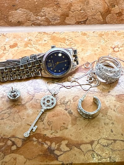 This close-up image displays a collection of silver-toned jewelry and a wristwatch laid out on a textured, light brown and beige surface resembling natural stone tiling or a countertop.
The primary subjects are a wristwatch, multiple rings, a necklace, and earrings. The wristwatch features a metallic link band and a dark blue face with small, luminous dots or crystals marking the hours, along with the Roman numeral "XII" at the 12 o'clock position. Faint text reading "SARAR SILVER" is visible on the watch face.
To the upper right of the watch are several silver-toned rings, appearing to be encrusted with small clear stones or diamonds, intertwined with a delicate silver chain necklace. Further right, a single silver-toned hoop earring, also adorned with stones, is visible. Below the necklace, a decorative key-shaped pendant, heavily encrusted with sparkling stones, hangs from the chain. Towards the bottom of the frame, a single stud earring, circular in shape and similarly decorated with stones, is present.
No activity is taking place; the items are stationary, seemingly placed down on the surface. The lighting suggests an indoor setting, possibly under artificial light or indirect daylight. The image does not contain any visual cues or specific elements that would indicate the location as "Going am Wilden Kaiser, Austria."