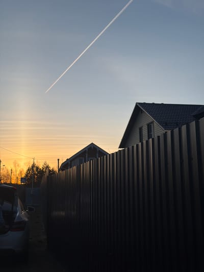 The image shows a scene in Воскресенские Усадьбы Кп, Russia, captured at what appears to be sunset or sunrise. The sky transitions from a pale blue at the top to a warm orange and yellow gradient near the horizon. A contrail is visible diagonally across the sky. The foreground is dominated by the dark silhouette of what seems to be a building or structure with horizontal siding and a peaked roof. Adjacent to this structure, there's another building with a tiled roof and what seems to be small windows. At the very bottom of the frame, there is a partial view of a vehicle with a visible taillight and side mirror. The horizon line features trees, possibly a small cluster, backlit by the sun, creating a dark silhouette. Power lines are also faintly visible against the bright sky. The image is oriented sideways.
