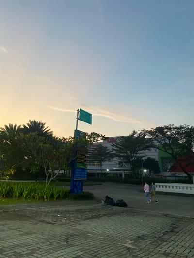 The image captures an outdoor scene at dusk in Mekar Bakti, Indonesia. In the foreground, a paved walkway made of interlocking rectangular bricks leads towards a street. To the left, a patch of green grass is bordered by rows of lush green plants. A tall signpost stands prominently, displaying directional arrows and text in various colors, indicating "Curug," "Cikupa," "ECO PLAZA," "SEKOLAH TARAKANITA," "the LEAF," "BELLE FLEUR," "WATER POINT," and "TAMAN TELAGA." Behind the signpost, palm trees and other foliage are visible, with a large building complex, possibly a commercial center or shopping mall, in the background. The building features multiple stories and appears to have signage with the letters "CGV" in red. Two individuals are walking away from the camera on the street. Several black trash bags are placed on the ground near the edge of the walkway. The sky above is a gradient of pale yellow near the horizon, transitioning to light blue with wispy clouds. Streetlights are visible, casting a soft glow on the scene.