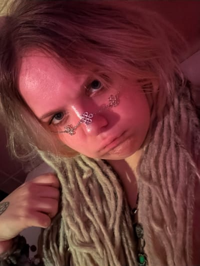 A close-up, low-angle, eye-level shot captures a young person's face illuminated by pink and red light, likely indoors in Vostochny District, Russia. The person has blonde hair styled in dreadlocks and is wearing silver facial jewelry consisting of chains and knot-like charms across their nose. Their skin appears flushed from the lighting, and they have blue eyes. The person's mouth is pursed, conveying a slightly pouty or contemplative expression. To the left, their left hand is clenched into a fist, with a tattoo visible on their forearm. The background is out of focus, but suggests an interior setting. The overall impression is one of intimate self-portraiture with a distinct aesthetic, possibly utilizing artificial lighting for effect.