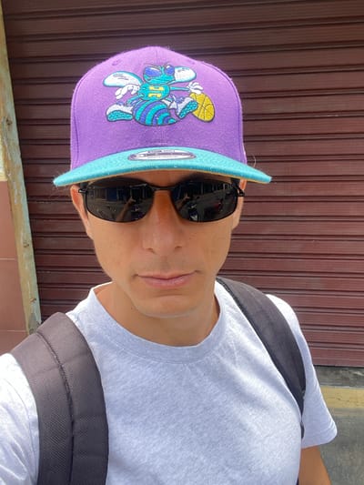 The image shows a close-up, portrait-style shot of a person wearing a purple baseball cap with a light blue brim. The cap features a cartoon character design with a basketball. The person is also wearing black sunglasses and a white t-shirt, with black straps of a backpack visible over their shoulders. The background consists of a dark brown, ribbed surface, possibly a door or wall, and a yellow and grey textured area. The image appears to be taken outdoors during the day, with natural lighting. The person's expression is neutral, and their face is partially obscured by sunglasses and the brim of the hat. The overall mood is casual. No discernible text is visible.