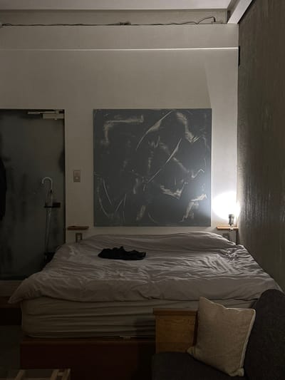 The scene depicts a dimly lit interior room in Suginami, Japan, primarily focused on a bed and its immediate surroundings. A large bed, covered in light grey sheets and a duvet, occupies the central background. A pair of dark athletic shoes rests on the duvet. Above the bed, a large, rectangular abstract artwork with a dark grey background and lighter, sweeping strokes is mounted on a light-colored wall.
To the left of the bed, a tall, industrial-style grey door is visible, featuring a door closer mechanism at the top and a light switch or outlet on the wall beside it. A transparent umbrella hangs from a stand positioned near the door's lower section, and a dark garment is suspended from a hook further left.
To the right of the bed, a small, wooden wall-mounted shelf supports a metallic, wall-mounted light fixture emitting a bright, focused beam onto the wall and bed. The adjacent wall on the far right has a textured, possibly concrete or rough finish. In the bottom right foreground, a section of a dark grey sofa with a light-colored, textured cushion is visible, alongside a light-colored wooden side table. Overhead, exposed wiring or conduit runs along the ceiling line. No individuals are present.