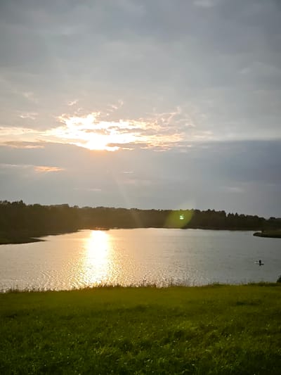 This media file captures a serene lake scene at sunset in Novosinkovo, Russia. The sun, partially obscured by clouds, casts a golden, shimmering reflection across the water. The sky is a mix of gray clouds and patches of sunlight, creating a dramatic and atmospheric effect. The foreground features a lush, green grassy hill.
The lake is bordered by a dense, dark treeline, suggesting a natural, undeveloped landscape. In the distance, a lone figure is visible on the water, possibly on a paddleboard or kayak, adding a touch of human presence to the vastness of the scene. The overall mood is peaceful and tranquil, evoking a sense of calm and natural beauty. The time of day is clearly sunset, given the warm light and the position of the sun. The weather appears to be partly cloudy, with the sun breaking through to illuminate the water. There is no visible text in the image.