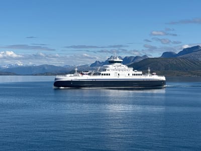 A wide shot captures a white ferry with a dark blue stripe navigating through calm, deep blue water under a bright, partly cloudy sky. The ferry, identified by text on its side, is moving from left to right, leaving a visible wake. In the background, a range of mountains with sparse vegetation and snow-capped peaks stretches across the horizon. The water's surface is rippled, reflecting the sunlight. The scene appears to be a scenic view of the water body near Lødingen, Norway.